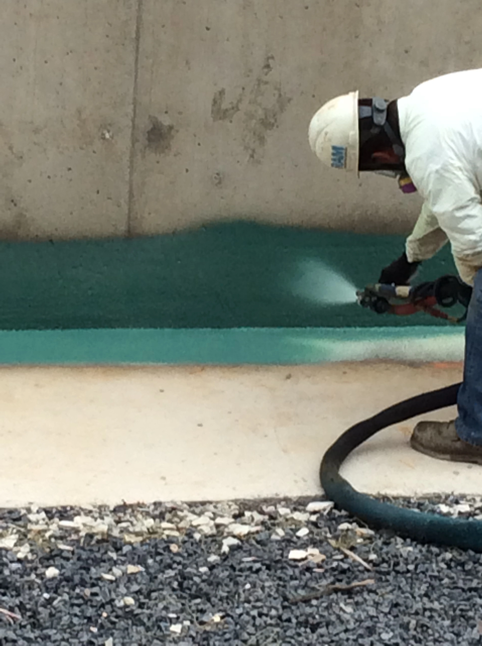 An image of a construction worker using a sprayer to apply a green coating to a bridge.